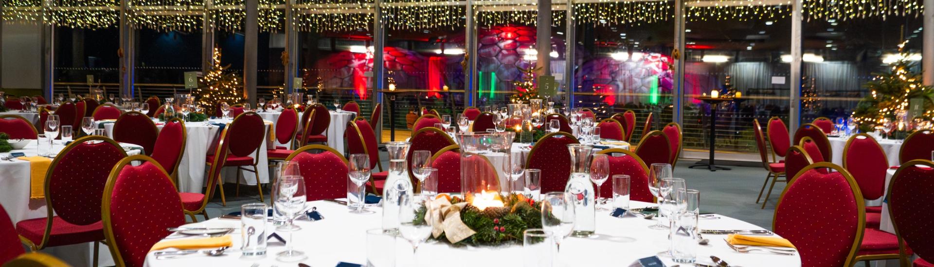 Christmas party tables set up with white tablecloths and red seating. There are twinkling fairy lights in the background.